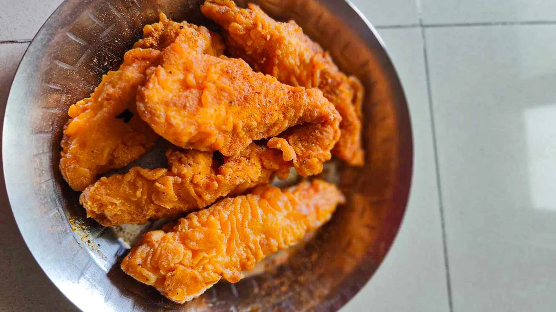 Battered and fried chicken pieces in a metal bowl on a tiled floor.